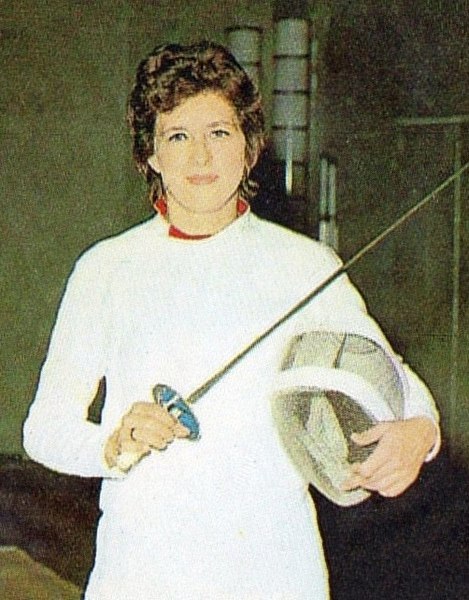 An old photo of Ujlaky-Rejto Ildiko holding a fencing foil and a helmet. She is in a white fencing jumpsuit.