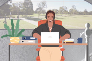 A captioner sitting at a desk with headphones and a microphone on. They are also smiling and typing on a laptop. There are books on their desk and a cup of tea next to them. There is a window behind them with a park and trees visible.