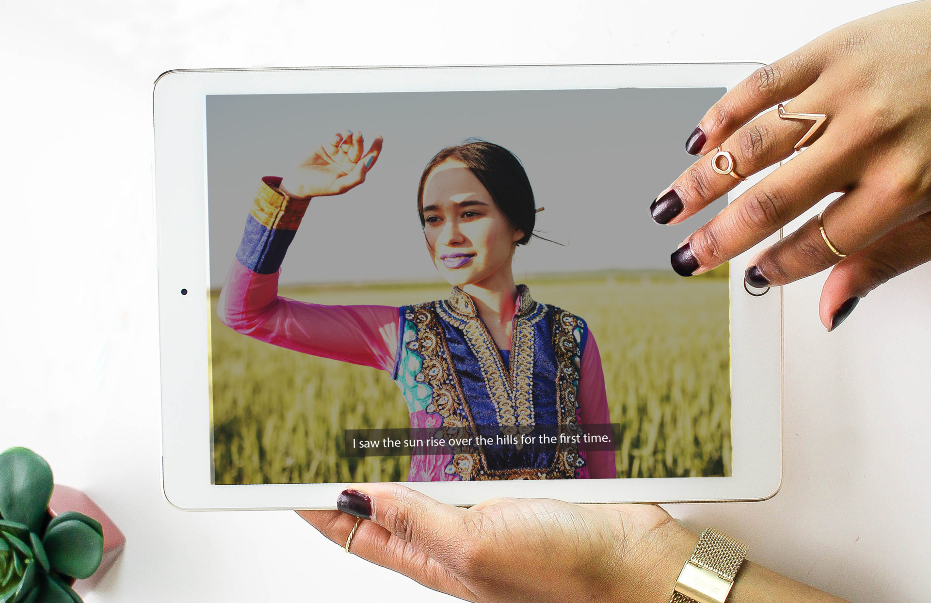 A person using a tablet device. The device screen pictures a young woman walking in a field. She is dressed in a colorful dress and a subtitle at the bottom of the screen reads: "I saw the sun rise over the hills for the first time."