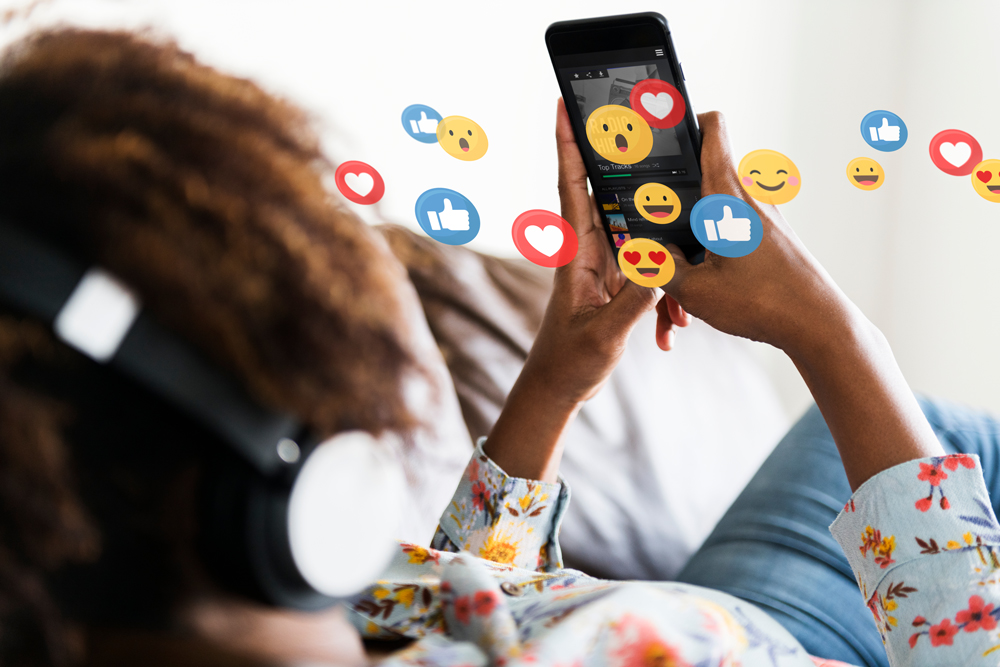 A person with headphones on is lying on a couch looking at their phone. A music app is playing on their phone and emojis are popping out of their screen.